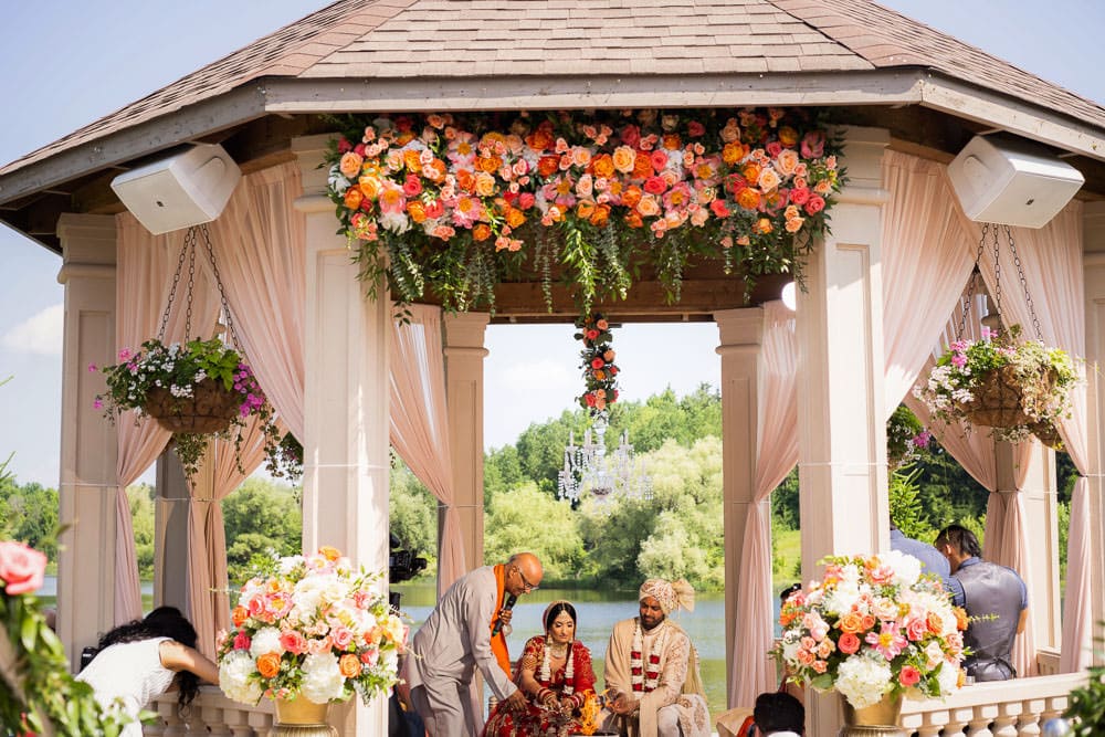 “Indian bride and groom standing outdoors near a historical monument, dressed in traditional red lehenga and cream sherwani, smiling and holding hands in natural sunlight.”