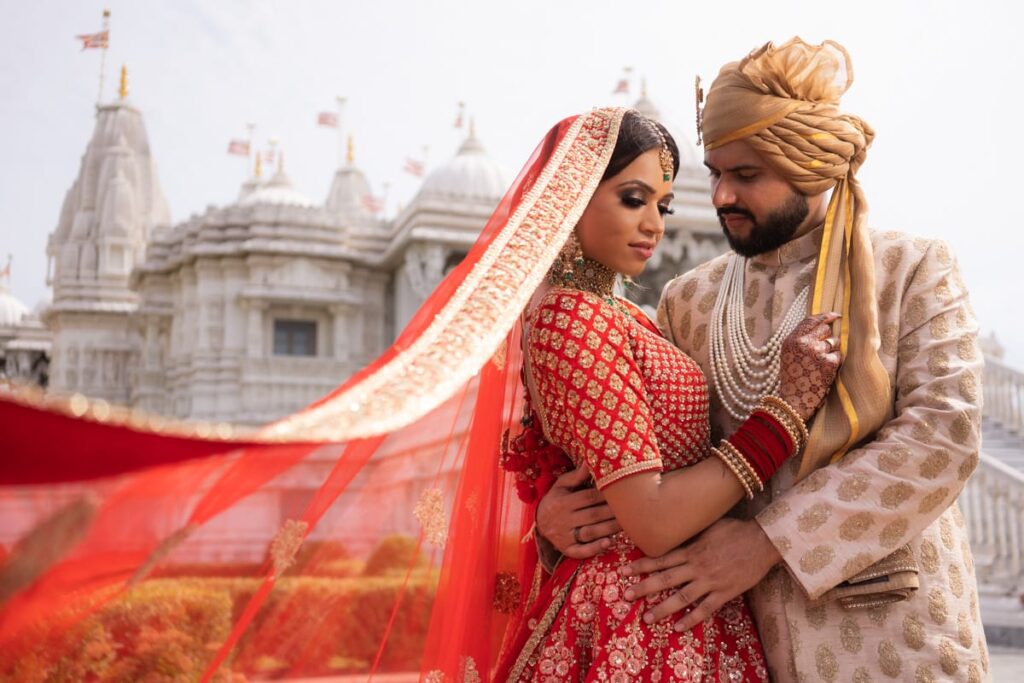 “Indian Sikh bride and groom holding hands in a garden during golden hour, dressed in red bridal lehenga and cream sherwani with warm outdoor lighting.”
